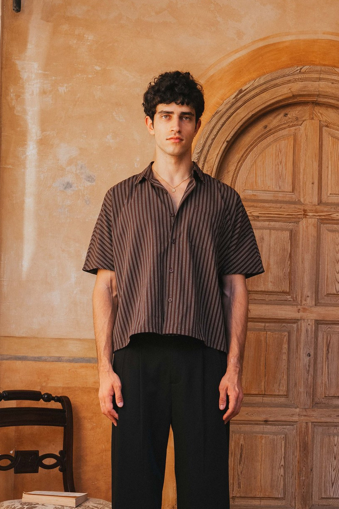 Man wearing a striped shirt and black pants standing in front of a wooden door.