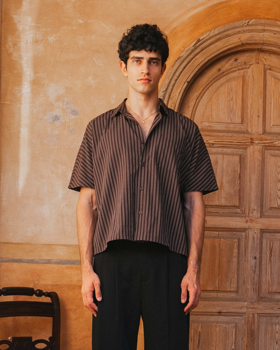 Man wearing a striped shirt and black pants standing in front of a wooden door.