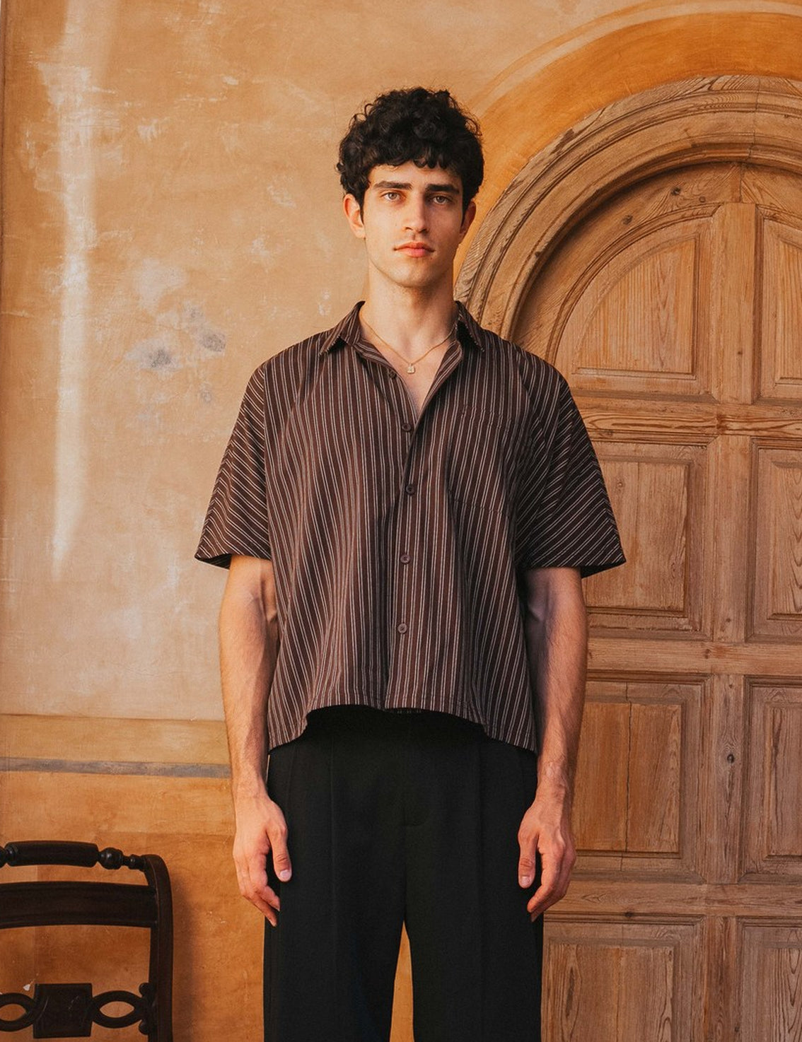 Man wearing a striped shirt and black pants standing in front of a wooden door.