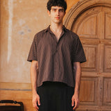 Man wearing a striped shirt and black pants standing in front of a wooden door.