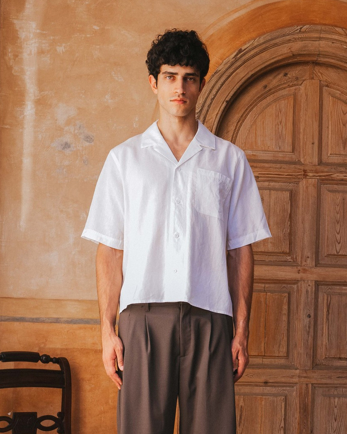 Man wearing a white shirt and brown pants standing in front of a wooden door.