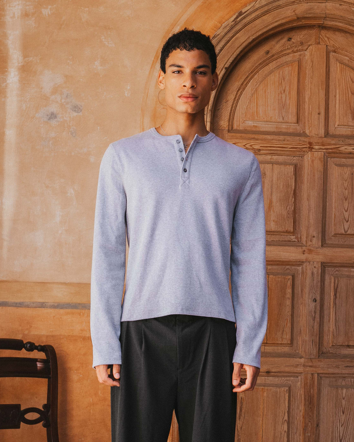 Man wearing a light blue long-sleeve shirt and dark pants standing in front of a wooden door.