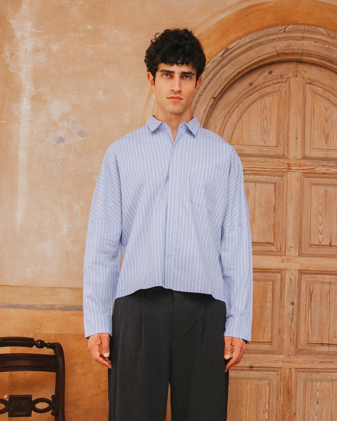 Man wearing a light blue striped shirt and dark pants standing in front of a wooden door.
