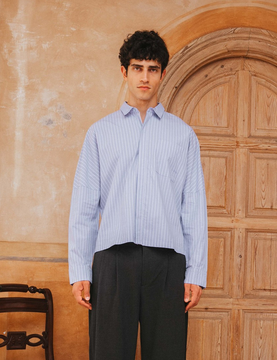 Man wearing a light blue striped shirt and dark pants standing in front of a wooden door.