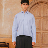 Man wearing a light blue striped shirt and dark pants standing in front of a wooden door.
