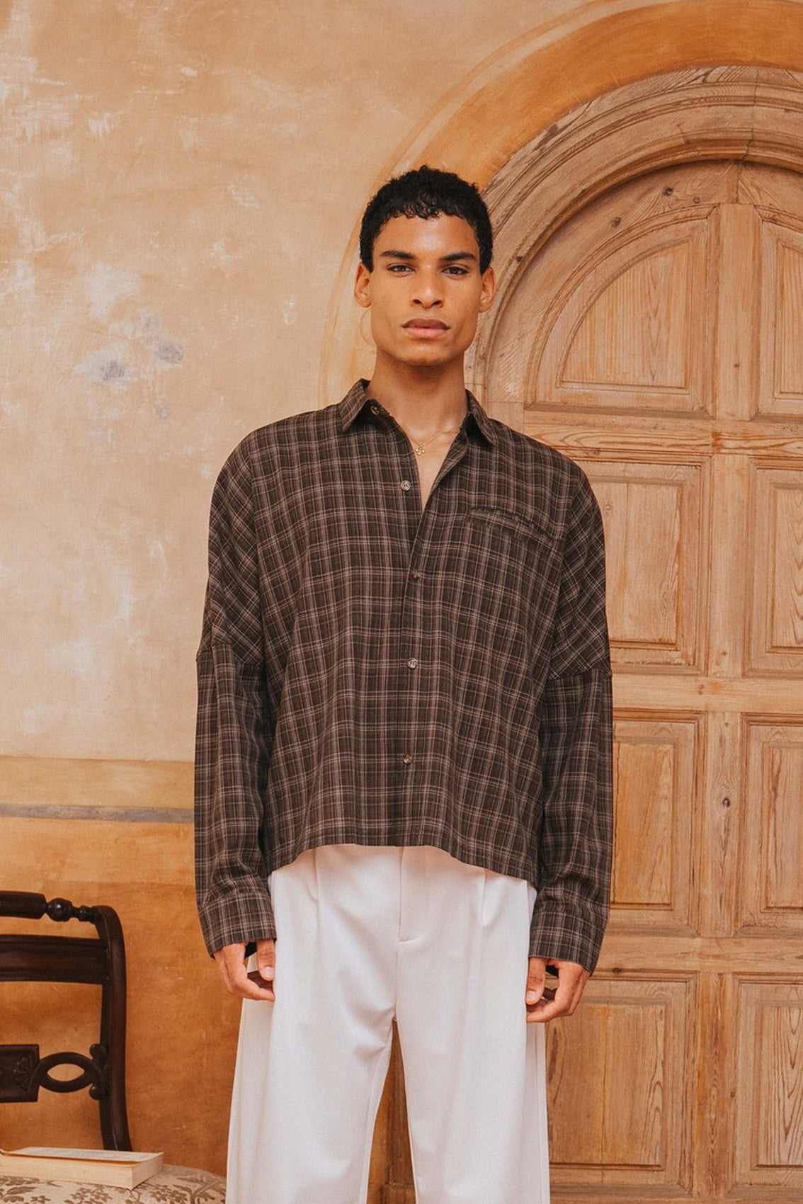 Man wearing a plaid shirt and white pants standing in front of a wooden door.